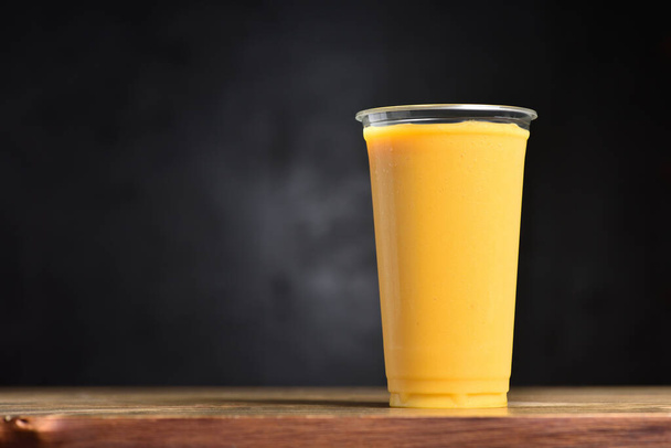Fruit smoothie in a plastic glass. Summer drink of mango, banana, ice and juice on a black background with copy space - Φωτογραφία, εικόνα