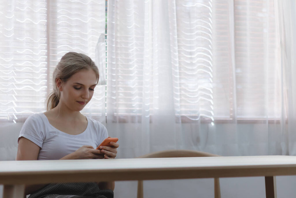 Młoda uśmiechnięta biała kobieta trzymająca smartfona za pomocą smsów do kochanka w domu. Dziewczyna korzystająca z telefonu komórkowego do zakupów online za pośrednictwem strony internetowej lub aplikacji. Koncepcja stylu życia i technologii - Zdjęcie, obraz