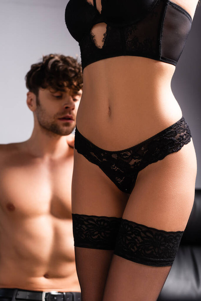Selective focus of sensual woman in lingerie and stockings standing near shirtless boyfriend on grey - Foto, imagen