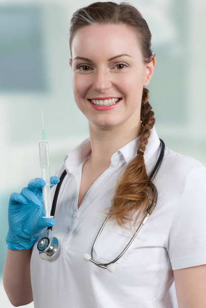 young female doctor or doctor's assistant with a pulled up syringe in front of a clinic room - Foto, Bild