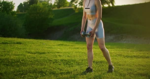 スポーツウェアを着た若い女性は、日没時に公園で自然界のダンベルで前進します。うまくいった。朝や日没時に美しい体に取り組む - 映像、動画