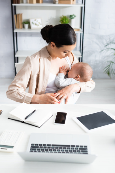 mother breastfeeding cute infant son near gadgets on table  - Photo, Image
