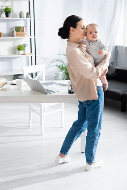 attractive mother standing and holding in arms cute infant son in baby romper near gadgets on table  - Φωτογραφία, εικόνα