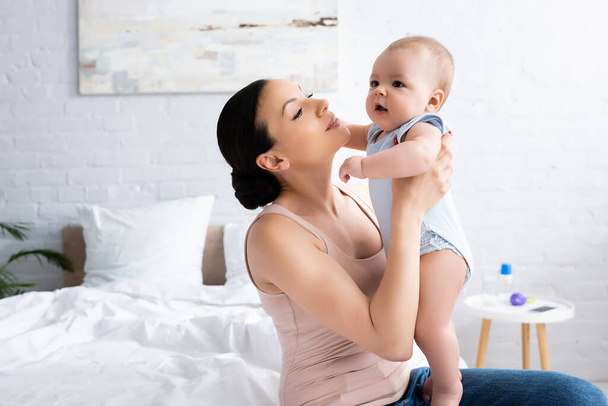 beautiful mother looking at cute infant son in bedroom - Foto, Imagem