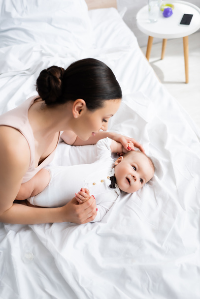 Mutter berührt entzückenden Sohn im Babystrampler mit Fliege auf Bett liegend - Foto, Bild