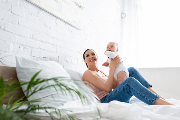 selective focus of happy mother in jeans sitting on bed and holding in arms cute infant son  - Φωτογραφία, εικόνα