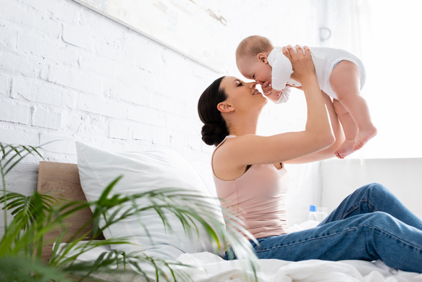 ベッドに座って腕の中で抱っこするジーンズ姿の明るい母親の選択的焦点かわいい幼児の息子  - 写真・画像