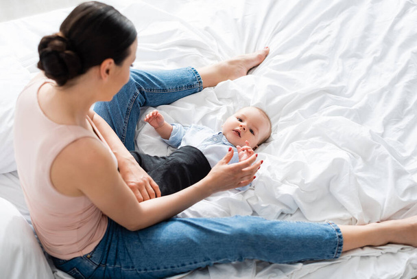 overhead view of mother in jeans sitting on bed and touching cute infant son  - Photo, Image