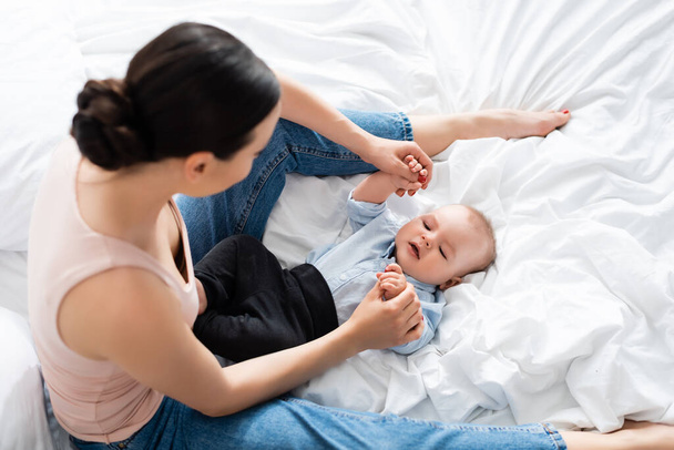 vista aerea di madre in jeans di jeans di jeans seduti sul letto e toccando le mani del figlio neonato  - Foto, immagini