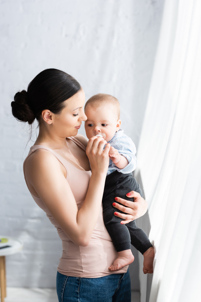 beautiful mother holding in arms barefoot infant son in baby clothing  - Photo, image