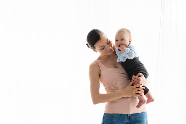 glimlachende moeder houden in armen blootsvoets zuigeling zoon in baby kleding  - Foto, afbeelding
