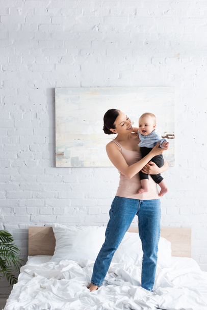 happy mother holding in arms happy barefoot infant son in baby clothing and standing on bed - Photo, Image