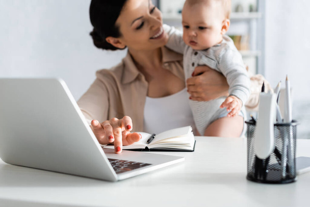 foco seletivo de freelancer feliz segurando em braços filho bebê e usando laptop
  - Foto, Imagem
