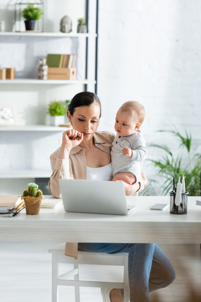 madre sosteniendo en brazos hijo bebé y mirando a la computadora portátil en casa
  - Foto, Imagen