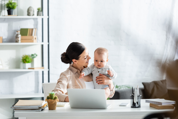 selective focus of happy mother looking at infant son near laptop on table - Photo, Image