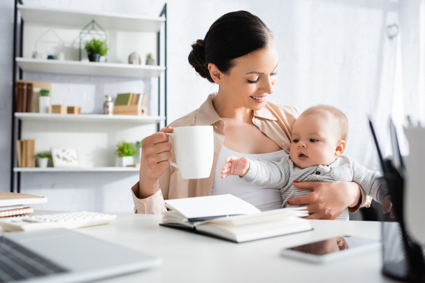 Розбірливо сфокусована на щасливій жінці, яка тримає чашку біля гарненького немовлячого сина і гаджетів.  - Фото, зображення