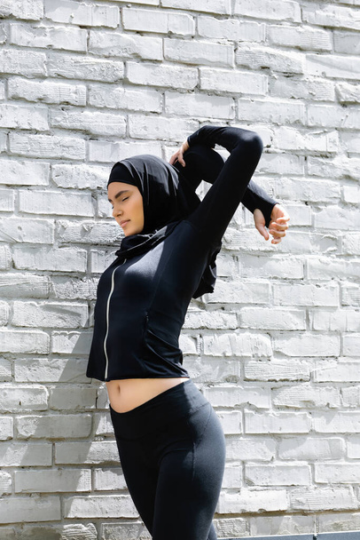 attractive muslim woman with closed eyes stretching near brick wall  - Photo, Image