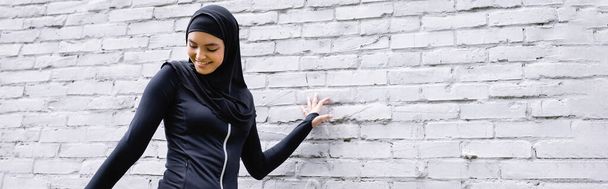 панорамный снимок привлекательной мусульманки, улыбающейся у кирпичной стены
  - Фото, изображение