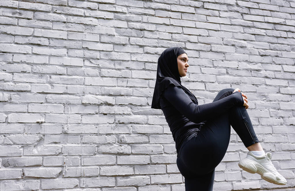 low angle view of attractive muslim sportswoman exercising near brick wall  - Фото, изображение