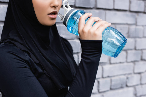 abgeschnittene Ansicht einer jungen muslimischen Sportlerin im Hijab, die draußen eine Sportflasche mit Wasser hält  - Foto, Bild