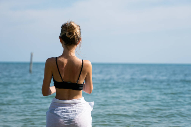 Young woman standing on a beach and enjoying the sun. Young woman in bikini looking at sea water on the beach of tropical resort. Rear view. Vacation and travel concept. - Zdjęcie, obraz