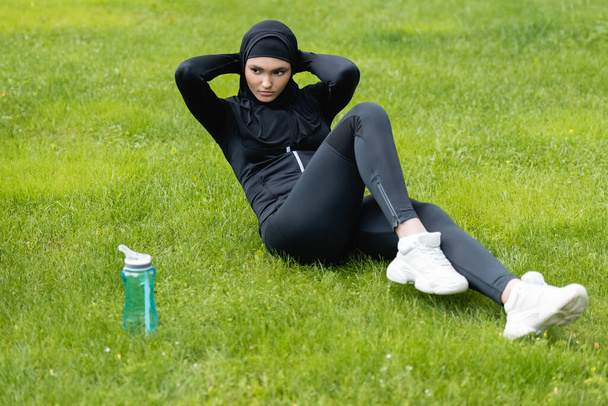 young muslim sportswoman in hijab exercising on grass near sports bottle with water  - Photo, Image