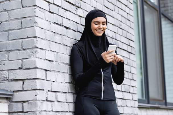 mujer musulmana feliz usando teléfono inteligente y de pie cerca de la pared de ladrillo
  - Foto, imagen