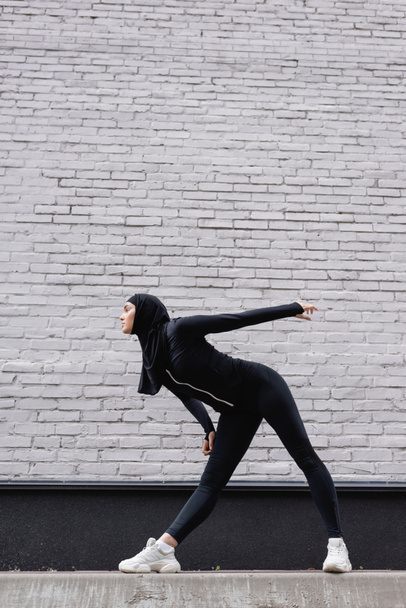 vista lateral da esportista árabe no hijab exercício perto da parede de tijolo
  - Foto, Imagem
