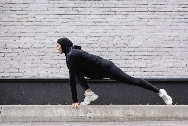 side view of arabian sportswoman in hijab stretching near brick wall  - Photo, Image