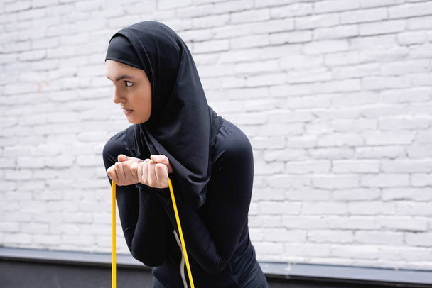 Arabisch meisje in hijab oefenen met weerstand band in de buurt van baksteen muur - Foto, afbeelding