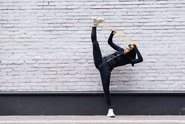 flexible arabian sportswoman in hijab exercising with resistance band near brick wall - Photo, Image