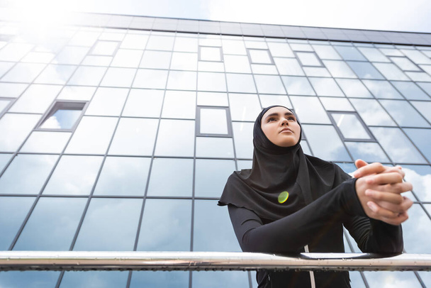 low angle view of muslim woman in hijab standing with clenched hands near modern building  - Fotó, kép
