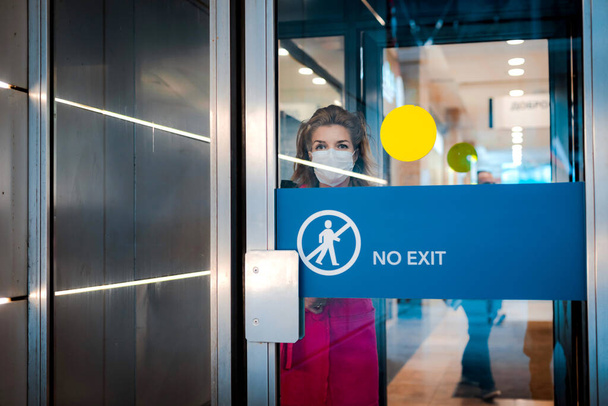 Stop infection. woman shows stop gesture through glass door there is no exit. girl wears a protective mask against infectious diseases flu in a public place. The concept of health, the quarantine. - Photo, Image