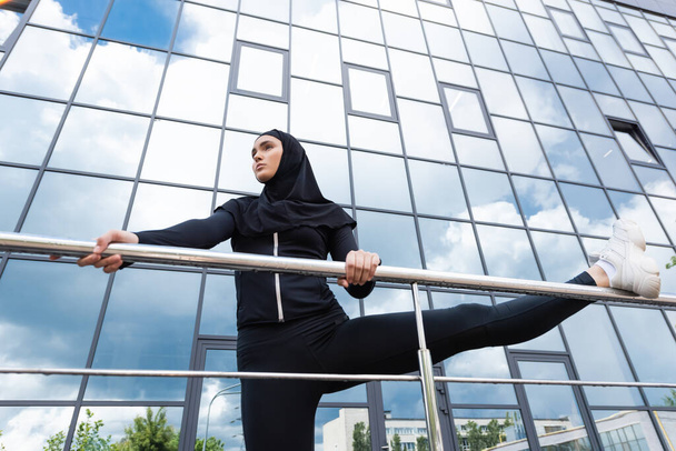 laag hoekzicht van Arabische vrouw in hijab houden leuning tijdens het sporten in de buurt van modern gebouw  - Foto, afbeelding