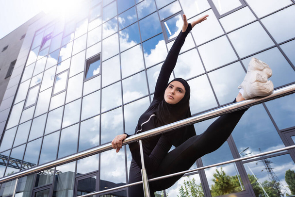Niedrigwinkel-Ansicht einer muslimischen Frau im Hijab, die beim Üben in der Nähe moderner Gebäude ein Geländer hält  - Foto, Bild