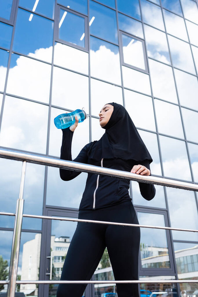 現代的な建物の近くにスポーツボトルを保持ヒジャーブ州のイスラムスポーツ女性の低角度ビュー  - 写真・画像