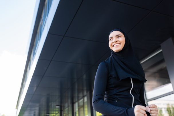 近くのビルで笑っているヒジャーブの幸せなイスラム教徒のスポーツ女性の低角度ビュー  - 写真・画像