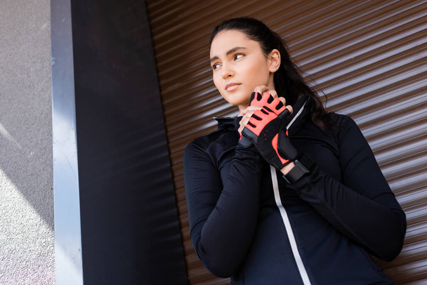 nízký úhel pohledu krásné mladé ženy dotýkající se sportovních rukavic a stojí venku - Fotografie, Obrázek