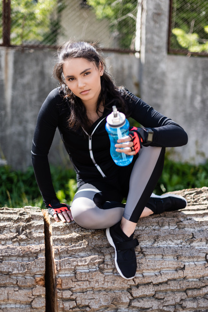 girl in sportswear holding sports bottle with water and sitting on tree trunk  - Foto, imagen