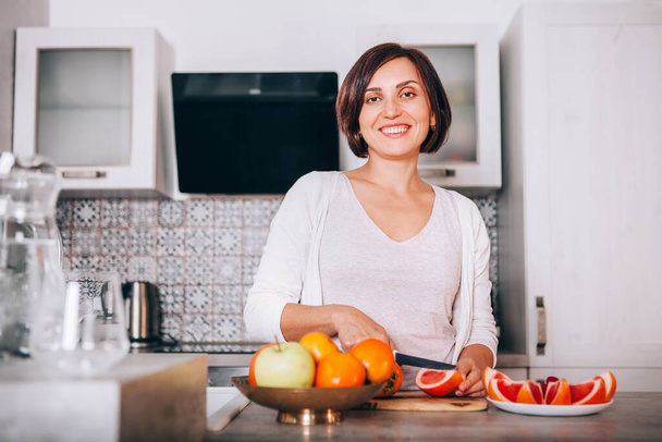 Beautiful young sincerely smiling female chopping grapefruit using a knife and cutting board in modern kitchen. Plenty of apples, grapefruits, kaki and oranges fruits, berries are on the tables.  - Foto, Imagen