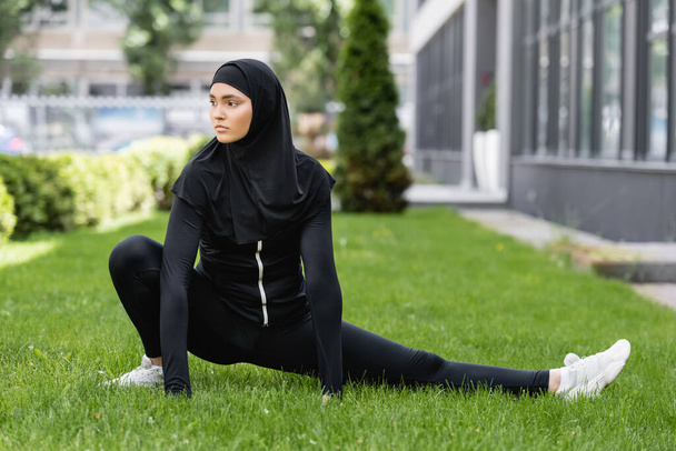 flexible arabian woman in hijab and sportswear stretching on green grass  - Photo, Image