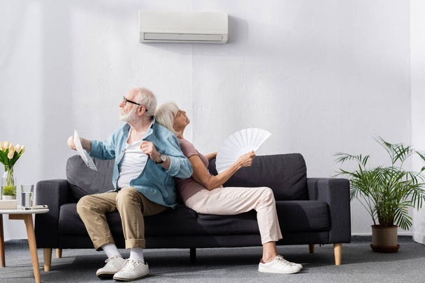 Senior man holding newspaper near tired wife with fan under air conditioner at home  - 写真・画像
