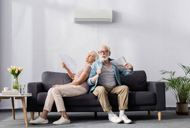 Mujer mayor sosteniendo ventilador mientras sufre de calor cerca de marido con periódico
  - Foto, Imagen