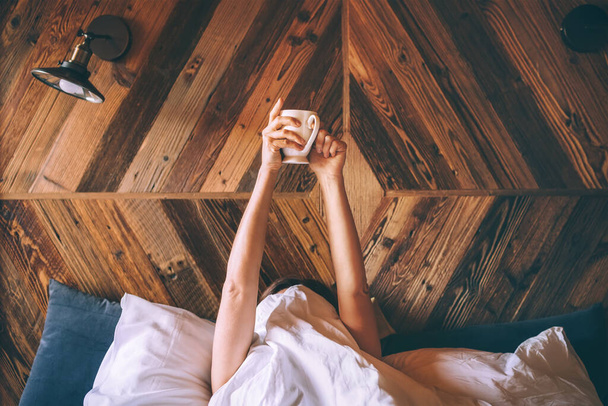Lazy female lying under the white blanket on the  linen bed and holding the fresh coffee cup in the early morning. Lazy day off and "coffee in bed"concept image. - Foto, Imagen