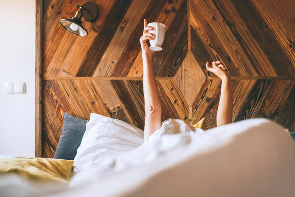 Lazy female lying under the white blanket on the  linen bed and holding the fresh coffee cup in the early morning. Lazy day off and "coffee in bed"concept image. - Foto, Bild