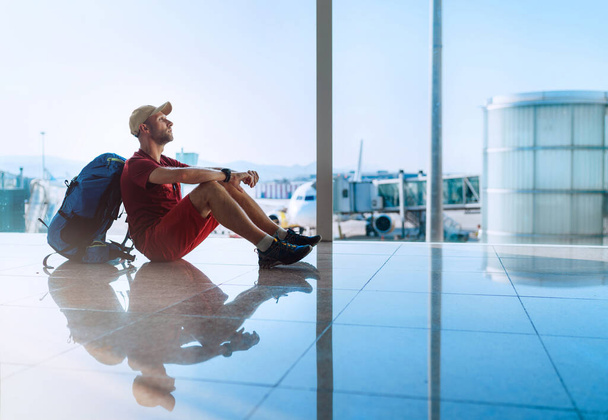 Alone backpacker traveller sitting on the airport terminal floor and waiting for boarding at aircraft which prepared for flight behind a window - 写真・画像