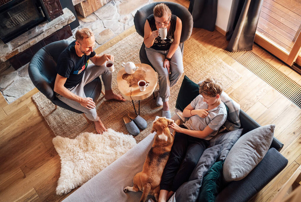 Cozy family tea time. Father, mother and son  at the home living room. Boy lying in comfortable sofa and  stroking their beagle dog and smiling. Peaceful family moments concept image. - Photo, Image