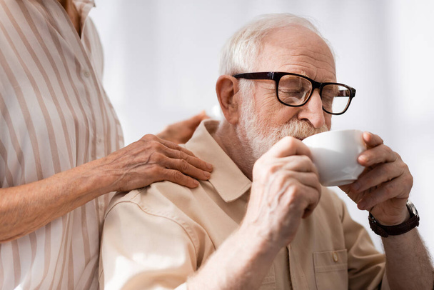 Selective focus of senior woman embracing husband drinking coffee at home  - Fotografie, Obrázek