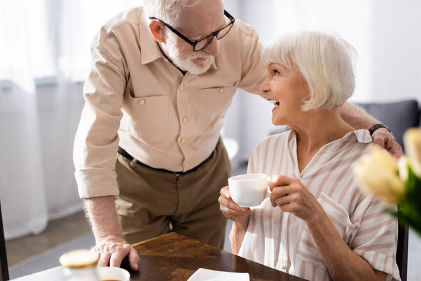 Selektywne skupienie się starszego mężczyzny przytulającego pozytywną żonę filiżanką kawy w kuchni  - Zdjęcie, obraz