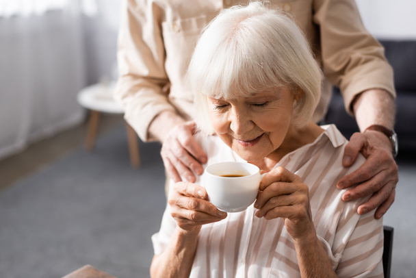 Selective focus of senior man embracing wife with cup of coffee at home  - Photo, Image